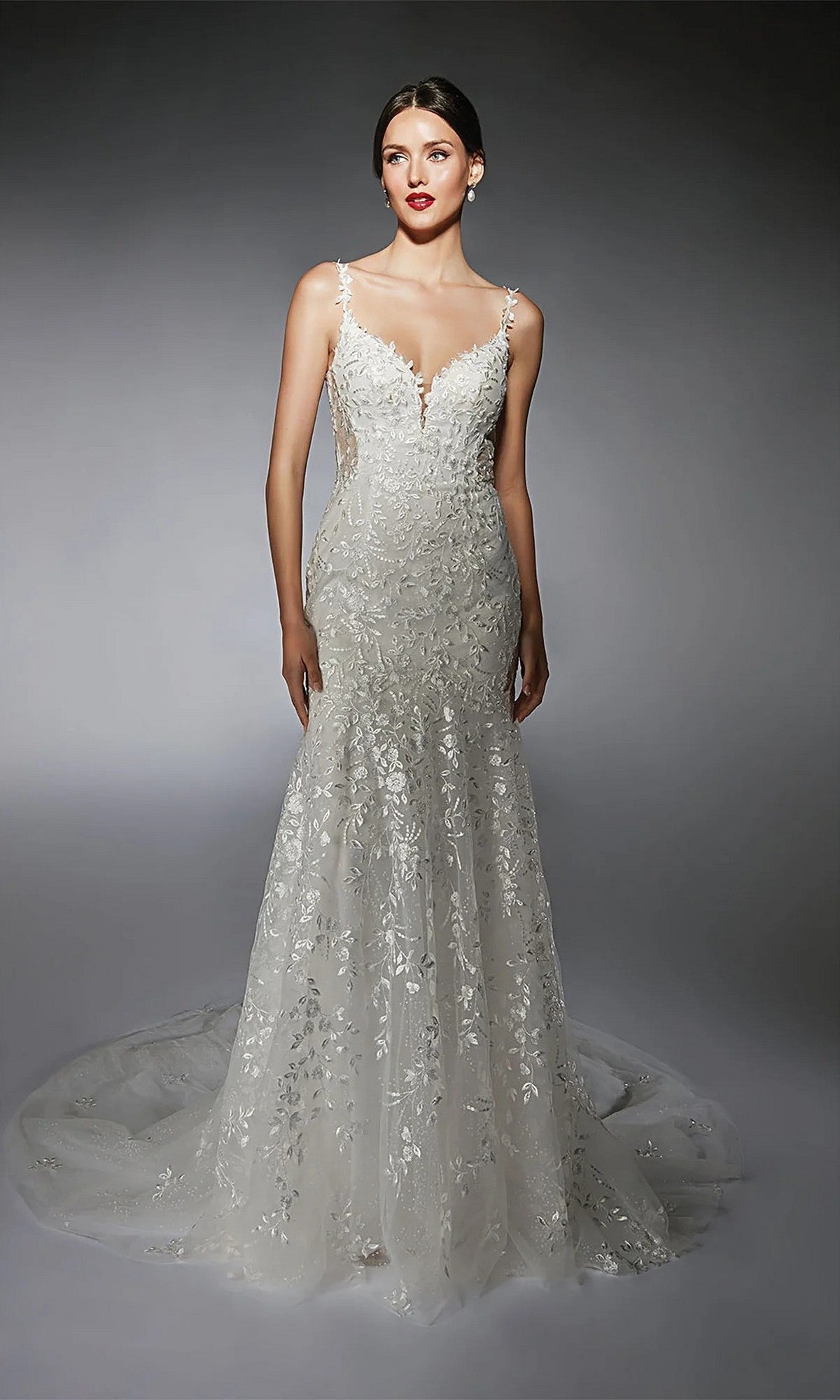 A woman stands against a gray gradient background wearing the fitted, sleeveless Long White Dress: Alyce 7120, featuring a floral lace overlay and a long, trailing train.